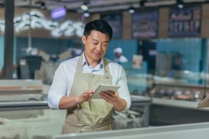 Asian grocery store manager salesman in apron using digital tablet counting goods in supermarket